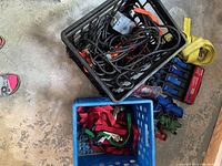 Photo from above showing two plastic crates; one containing black coiled extension cord and the other red ratchet straps; assorted ratchet straps of various colors and metal hooks laid out on floor.