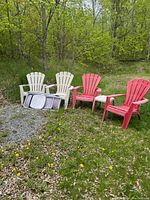 Photo showing four outdoor resin Adirondack chairs, two red and two cream, placed on grass in a wooded area. Two metal folding chairs stacked on the ground in front of the cream chairs.