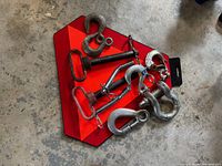 Photo showing a collection of metal hooks of various sizes and types, some with safety latches and clevis pins, all placed on top of a red and orange reflective slow moving vehicle hazard sign with a black handle.