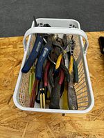 White plastic basket containing a variety of used hand tools including wrenches, screwdrivers, and pliers with mixed wear and rust present.