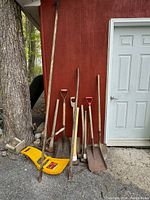 Full view of ten outdoor tools propped against a red wall and a door, including shovels, pick axe, maul, sledge hammers, folding shovel, and wood limb cutter.
