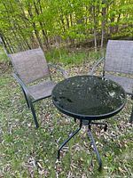 Small patio set shown outdoors on grass with trees in the background. Includes a small round glass top table and two mesh fabric chairs with metal frames.