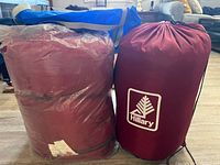 Two single sleeping bags, one in a red storage sack with Hillary logo and the other in a transparent plastic cover.