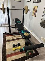 Adjustable bench with attached preacher curl pad and leg rollers; loaded barbell resting on rack; weight plates and dumbbells on floor