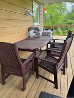 Five solid wood vertical slatted back chairs with armrests arranged around a rectangular slatted top patio table on a wooden deck. Cushions wrapped in plastic bags placed on top of the table.