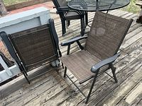 Two outdoor folding chairs shown on deck, brown mesh fabric seats and backs, black metal frames, black armrests, some wear visible