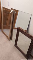 Three mirrors stacked leaning against a wall including one gold painted frame, one wooden frame, and one non-framed mirror.