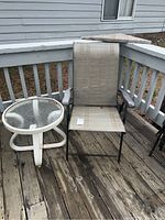 Photo of outdoor chair with beige mesh fabric seat and back, black metal frame, and white round side table with glass top on wooden deck