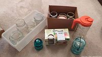 Photo showing assorted mason jars including three clear glass jars in a white plastic container, unopened box of Ball mason jars, boxed lids in a cardboard box, and Tang brand orange-lidded glass pitcher.
