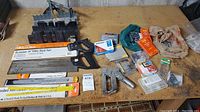 Photo showing miter boxes, three back saws including an Economy 14" Miter Back Saw, staple device, staples, and small hardware items arranged on a wooden bench.