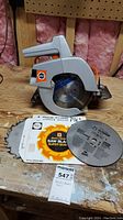 Front view of Black & Decker circular saw on wooden surface with two packaged saw blades laid in front.