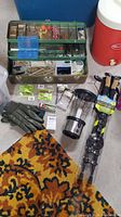 Photo showing green fishing tackle box with fishing baits, gloves, lantern, packaged fishing rod, cooler and beverage dispenser on basement floor with orange and black patterned mat under some items.
