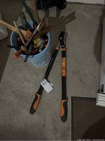 Full view of two Fiskars loppers with orange and black handles lying on concrete beside a blue bucket holding assorted tools.