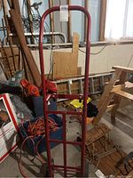 Red metal hand truck dolly standing upright in garage setting among various tools and equipment.