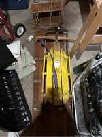 Photo showing old wooden yellow sled with metal runners and purple pull rope, resting on floor with another wooden toboggan underneath.