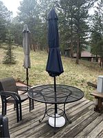 Photo of round metal mesh table with closed dark blue umbrella in white base on wooden deck outdoors.