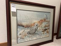 Framed print showing two snowy owls in a winter landscape, with a dark wood frame and matting, includes auction tag.