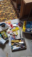Assortment of painting tools and supplies including brushes, rollers, trays, extension poles and small cans scattered on concrete floor near patterned carpet and wooden cabinetry.