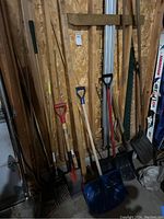 Photo showing multiple garden tools including shovels and rakes leaning against a plywood wall in a garage or storage area.