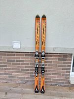 Full view of the pair of orange Atomic 9.22 snow skis with bindings against white wall and brick section.