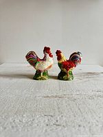 Pair of multicolored ceramic chicken salt and pepper shakers on fabric surface, showing front and side views