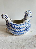 Side view of the large pottery bowl shaped like a rooster showing detailed blue painted leaves and feathers on a white speckled ceramic surface.