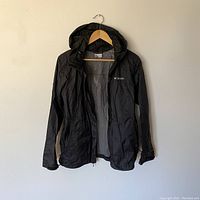 Front view of black lightweight Columbia windbreaker jacket hanging on a wooden hanger against a plain wall, showing hood, full front zipper, and Columbia logo on left chest.