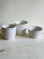 Three small white ceramic planter pots arranged on a light wooden surface, showing distinct shapes and textures.