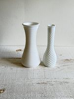 Photo showing two white milk glass vases side by side on a textured surface with neutral wall background, one vase taller with horizontal ribbed pattern, the other shorter with diamond pattern.