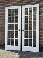 Two white French interior doors with glass panels placed upright against a brick wall, showing front view with visible glass grid panels and door knobs.