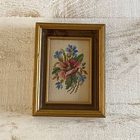 Frontal view of the vintage flower stitched artwork in its decorative wood and gold frame lying on a beige textured surface.