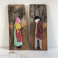 Two side profile carved wooden sculptures of an old woman and man, each mounted on a weathered reclaimed wooden shingle.