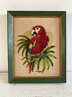 Front view of vintage handmade crewel embroidery wall hanging featuring a red parrot perched on green tropical leaves. In green wooden frame with gold trim.