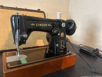 Side perspective of black antique Singer sewing machine mounted on polished wooden base, showing machine details such as needle area, foot pedal, and spools.