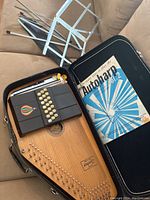 Full view of the autoharp inside a black hard case with instruction book visible and metal music stand in background