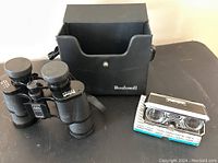 Bushnell binoculars with carrying case and Ensign folding opera glasses on black surface.