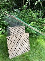 Metal fencing rod with attached green plastic fencing and two beige lattice panels leaning against green shrubs outdoors