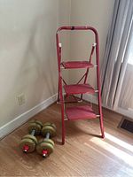 Photo showing red 3-step folding ladder with paint chipping and pair of dumbbells on wood floor by wall