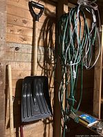 View of snow shovel with D-shaped handle, wooden shaft, and black scoop next to broom handle and several extension cords hanging on wood wall.