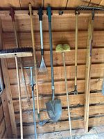 Photo of 9 gardening tools hanging on a wooden shed wall including a broom, 3 rakes, a hoe, 2 weed pullers, and a shovel.