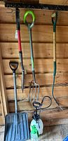 Photo showing seven gardening tools hanging on wooden shed wall including shovels, pitch fork, ergonomic hoe, snow shovel, and two weed sprayers.