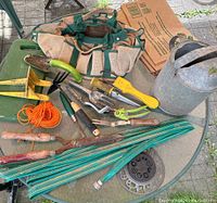 Photo of garden tools including antique watering can, garden hand tools, garden tool bag, garden hose, sprinkler, hose reel, orange twine and knee pad on glass table outdoors.