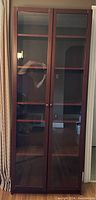 Front view of tall IKEA Billy bookcase with closed glass doors showing dark wood finish and visible reflection.