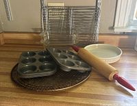 Photo showing five metal cooling racks stacked vertically, two rectangular glass Pyrex loaf pans, a white ceramic round quiche/flan pan, three metal muffin tins (two 6-cup and one 12-cup), a metal pizza tray, and a wooden rolling pin with red handles lying on a kitchen counter.