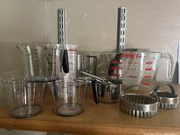 Photo showing multiple measuring cups and pitchers including Pyrex glass with red markings, stainless steel measuring cups, and Pampered Chef plastic measuring tools and cutters.
