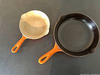 Two orange-handled cast iron frying pans with black interiors, smaller pan showing some residue, placed side by side