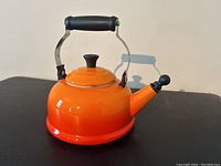 Red orange Le Creuset kettle with metal handle and black rubber grip, shown from the side.