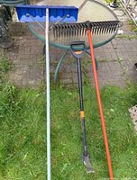 Photo of three garden tools including blue roof rake, green metal leaf rake, and Fiskars weeding tool.