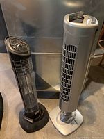 Photo showing two vertical tower appliances: a silver Ener-g+ heater tower with a control panel and remote on top, and a black cooling fan tower on a black base.