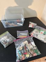 Lot displayed on a black surface with plastic storage container and several small zipper bags containing sewing notions including thread spools and other notions.